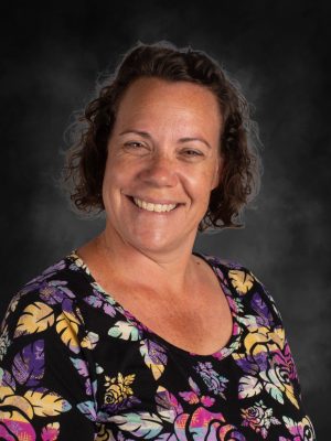 Mrs. Andrea Clausen smiling in a floral shirt against a dark background.